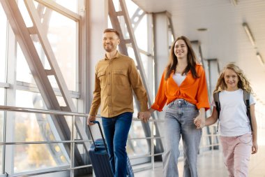 Küçük kızı olan mutlu bir beyaz aile havaalanında yürüyor, birlikte vakit geçiriyorlar, neşeli bir seyahat tarzı ve birliktelik sergiliyorlar.