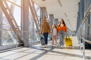 Mutlu Avrupalı aile havaalanında bavullarıyla yan yana yürüyor, el ele tutuşuyor ve gün ışığında gülümsüyor, neşeli bir bağ, yaşam tarzı ve tatil tatili neşesini simgeliyor.
