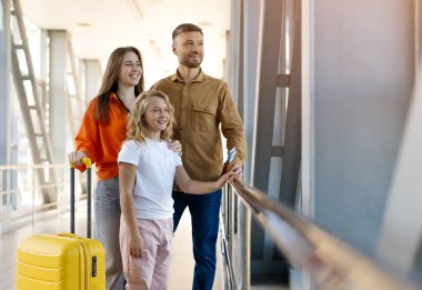 Beyaz bir aile havaalanında. Pasaportları ve bavullarıyla pencereden dışarı bakıyor. Birlikte gülümsüyor, neşeli bağları, yaşam tarzını ve yurtdışında seyahat mutluluğunu vurguluyor.