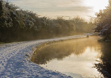 View along British rural countryside canal in winter with snow covered towpath and beautiful misty sunrise clipart