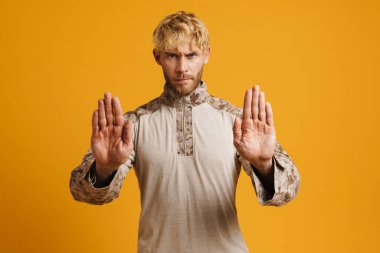 Üniformalı Avrupalı asker sarı arka planda izole edilmiş kameraya dur işareti yapıyor.