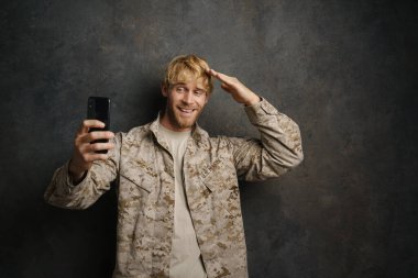 Beyaz asker, siyah arka planda izole edilmiş cep telefonuyla selfie çekerken selam veriyor.