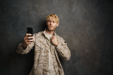 Üniformalı beyaz asker siyah arka plandan izole edilmiş cep telefonuyla selfie çekiyor.