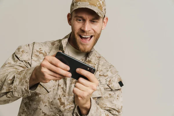 stock image White military man wearing uniform playing online game on cellphone isolated over white background