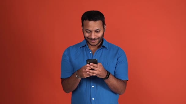 快乐的印度人穿着蓝色T恤衫在橙色工作室里用手机打字 — 图库视频影像