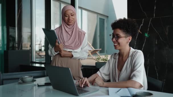 两名笑着的多族裔穆斯林妇女和非洲妇女在办公室里用笔记本电脑聊天和工作 — 图库视频影像