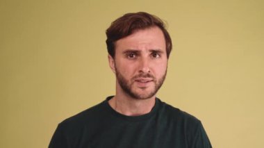 Tişört giyen endişeli esmer genç adam sarı stüdyoda kameraya seviniyor.