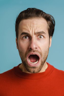Kırmızı kazak giyen genç adam mavi arka planda kameraya bağırıyor.