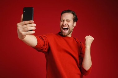 Genç adam kırmızı arka planda izole edilmiş cep telefonuyla selfie çekerken kazanan hareketi yapıyor.
