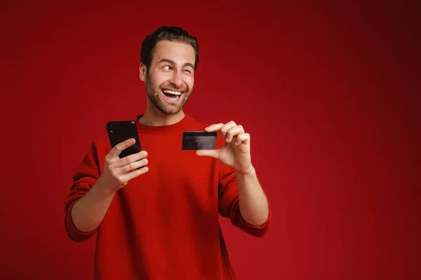 在使用红色背景隔离的手机和信用卡时 年轻的白发男子眨眼了 — 图库照片