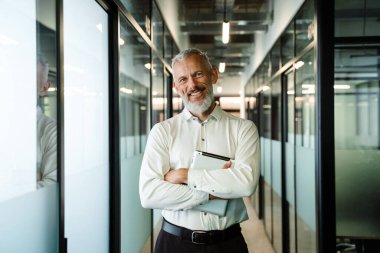 Ofiste tablet bilgisayarla poz verirken kameraya gülümseyen gri beyaz adam.