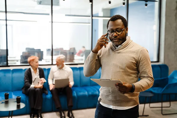 同僚とオフィスで仕事をしながら携帯電話で話す髭黒の男 — ストック写真