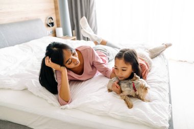 Asian mother and daughter smiling while lying on bed with their dog at home