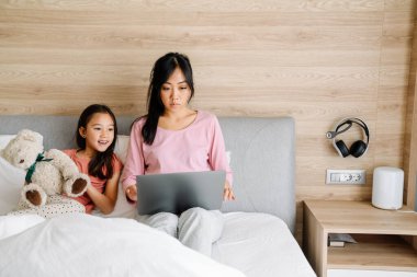 Asian mother and daughter using laptop while sitting on bed at home