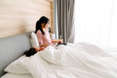 Asian woman using laptop while her daughter sleeping on bed at home