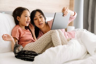 Asian mother and daughter using tablet computer while sitting on bed at home