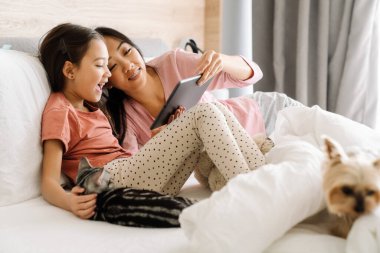 Asian mother and daughter using tablet computer while sitting on bed at home