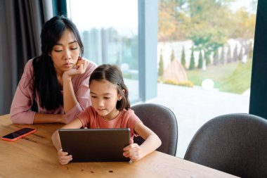 Asian woman and her daughter using tablet computer while sitting at table in home
