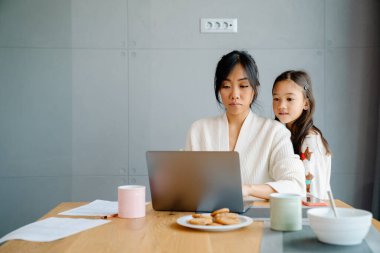 Asian woman working with laptop while her daughter doing homework at home