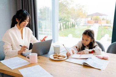 Asian woman working with laptop while her daughter doing homework at home