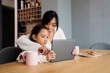 Asian woman and her daughter using tablet computer while sitting at table in home