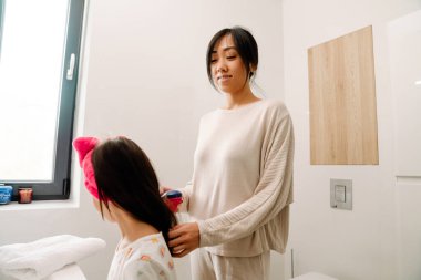 Asian mother combing her daughter's hair in bathroom at home