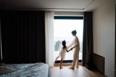 Asian woman and her daughter looking out window together at home