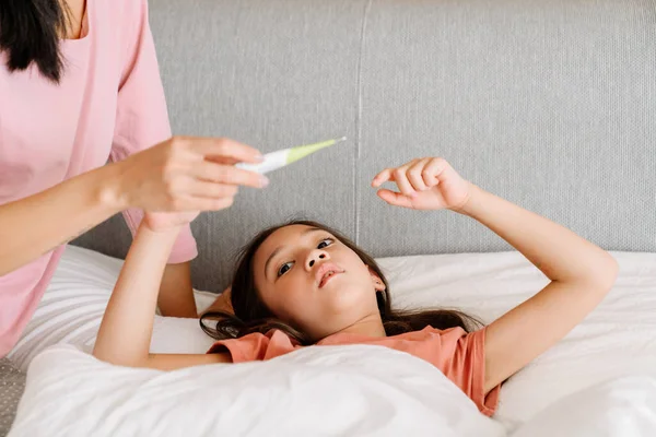 Asian girl with flu measuring temperature while lying on bed at home