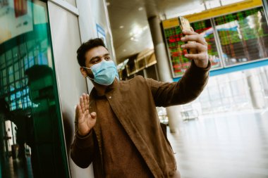 Yüz maskesi takan Hintli bir adam kapalı bir tren istasyonunda cep telefonundan selfie çekiyor.