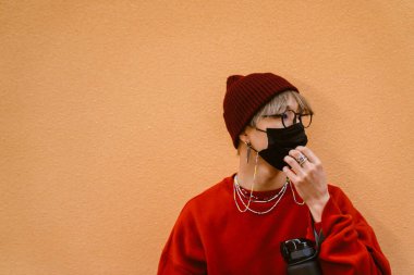 Suratı maskeli ve gözlüklü Asyalı çocuk dışarıda duvarın yanında duruyor.
