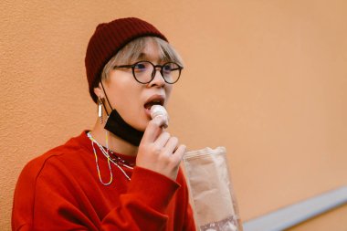 Gözlüklü Asyalı çocuk dışarıda duvarda dururken kurabiye yiyor.