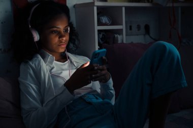 Telefonla müzik dinleyen ve evdeki karanlık odada video izleyen Afrikalı bir genç kız.