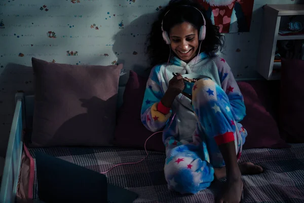 アフリカの幸せな笑顔女の子でパジャマとヘッドフォン食べるシリアルとともにミルクオンベッド ホーム — ストック写真