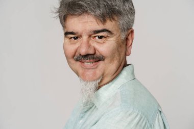 Bıyıklı, sakallı, beyaz arka planda kameraya gülümseyen yaşlı gri adam.