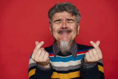 Gri sakallı, yaşlı bir adam, yüzünü buruşturuyor ve parmaklarını kırmızı arka planda soyutluyor.