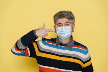 Olgun gri adam sarı arka planda izole edilmiş maskesini işaret ediyor.