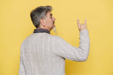 Gri sakallı olgun bir adam boynuz hareketi yapıyor ve sarı arka planda izole edilmiş bir kenara bakıyor.