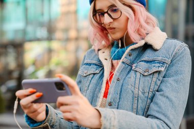 Gözlüklü genç kadın şehir caddesinde dikilirken cep telefonu kullanıyor.