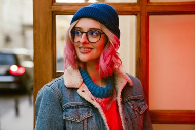 Gözlüklü genç bir kadın şehir caddesinde dikilirken gülümsüyor.