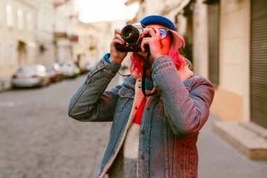 Şehir caddesinde yürürken retro kamerada fotoğraf çeken genç bir kadın.
