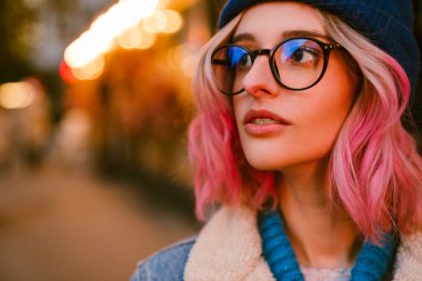 Gözlüklü genç kadın şehir caddesinde dikilirken yana bakıyor.