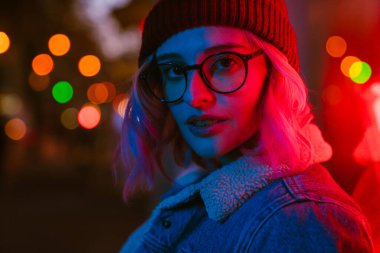 Gözlüklü genç beyaz kadın şehir caddesinde kameraya bakıyor.