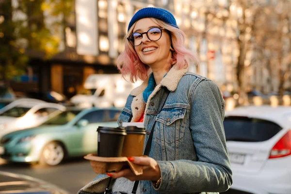 stock image Young woman in eyeglasses laughing and drinking coffee while walking on city street