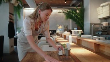 Gülümseyen sarışın bayan garson kafede masaları siliyor.