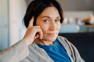 Beyaz, olgun bir kadın evdeki koltukta otururken kameraya bakıp gülümsüyor.