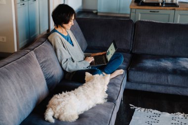 Beyaz, olgun bir kadın evinde köpeğiyle otururken dizüstü bilgisayar kullanıyor.