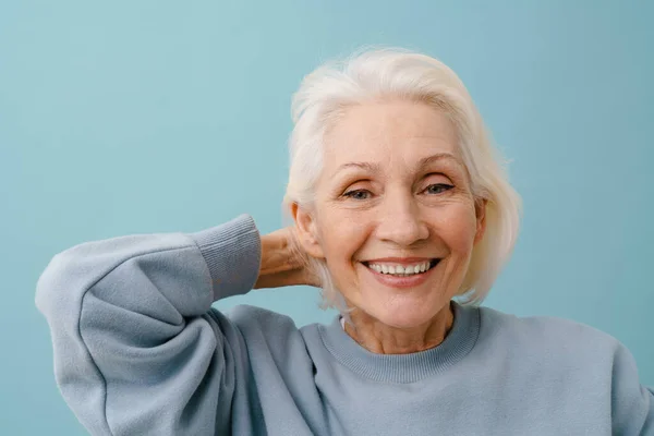穿着蓝色套头衫的漂亮而快乐的老年妇女把头发调整到孤立的蓝色背景上 — 图库照片