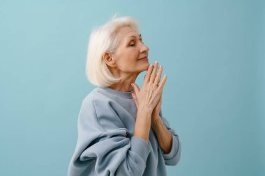 Mavi kazaklı, güzel, yaşlı bir kadın. Gözleri kapalı, elleri katlanmış, mavi arka planda.