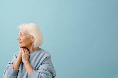 Mavi kazaklı, güzel, yaşlı bir kadın. Gözleri kapalı, elleri katlanmış, mavi arka planda.