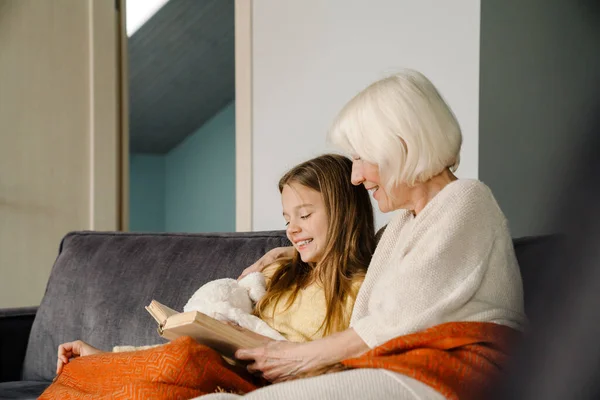 Branco Menina Leitura Livro Com Ela Avó Enquanto Sentado Sofá — Fotografia de Stock